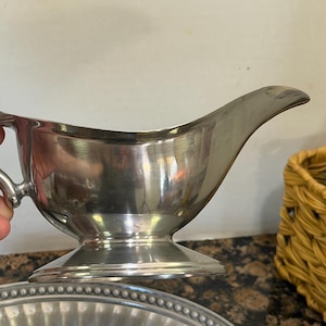 May include: A polished silver-tone metal gravy boat featuring a curved handle, a wide pouring spout, and a circular pedestal base. The vessel has a smooth, reflective surface and a classic silhouette designed for serving sauces or gravies.
