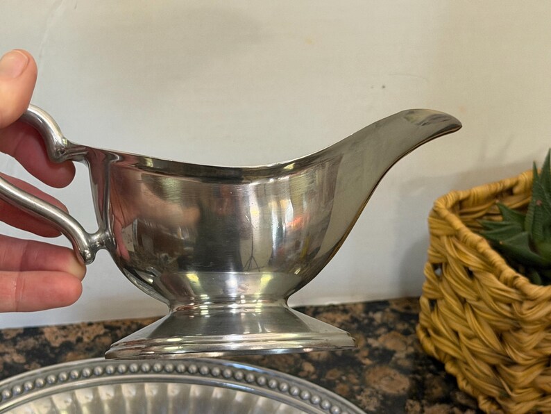 May include: A polished silver-tone metal gravy boat featuring a curved handle, a wide pouring spout, and a circular pedestal base. The vessel has a smooth, reflective surface and a classic silhouette designed for serving sauces or gravies.