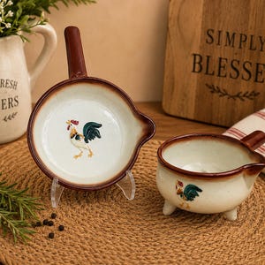 May include: Two ceramic kitchen vessels with a cream-colored base and brown rims. One is a shallow pan-shaped dish with a handle, and the other is a small three-legged bowl with a handle and spout. Both feature a painted rooster illustration.