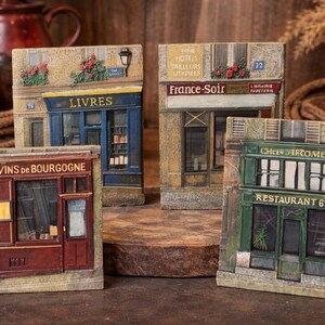 May include: A collection of three rectangular decorative plaques featuring detailed relief models of French storefronts. The plaques depict a red wine shop labeled Vins de Bourgogne, a blue bookstore labeled Livres, and a green restaurant labeled Chez Jerome.