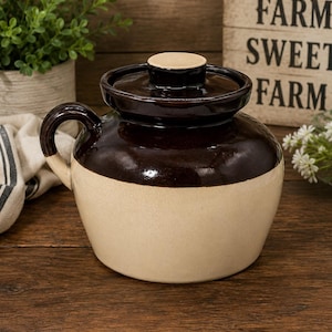 May include: A ceramic crock featuring a two-tone design with a dark brown glazed upper section and a cream-colored lower body. The vessel includes a matching dark brown lid with a central knob handle and a single side handle for gripping.