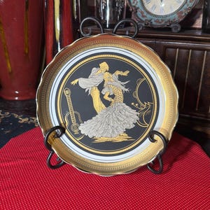 May include: Decorative plate with a black center featuring a gold and white illustration of a dancing couple and a guitar. The plate has a gold rim and is displayed on a black metal stand. The plate is on a red surface.
