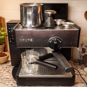 May include: A black Krups espresso machine with a stainless steel top surface, a front-facing control dial, and a portafilter handle. A clear glass measuring pitcher with a black handle sits on the drip tray, while a metal milk frothing pitcher rests on top.