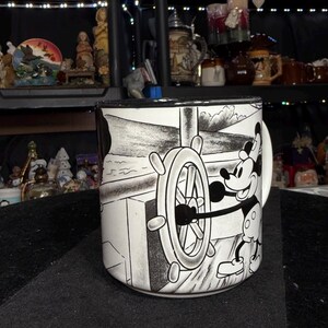 May include: A black and white ceramic mug featuring Mickey Mouse at the helm of a ship. The mug has a black rim and handle. The design depicts Mickey Mouse steering a ship's wheel with a nautical background.