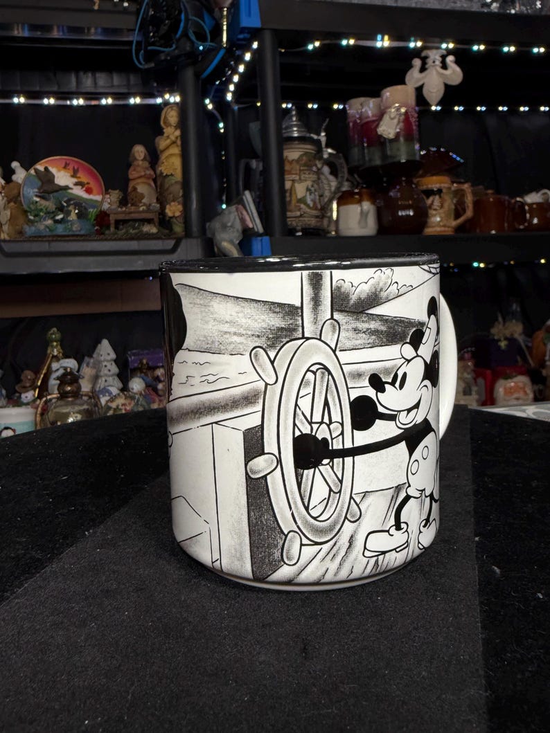 May include: A black and white ceramic mug featuring Mickey Mouse at the helm of a ship. The mug has a black rim and handle. The design depicts Mickey Mouse steering a ship's wheel with a nautical background.