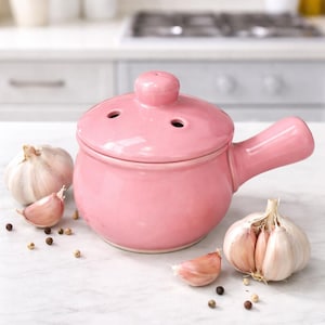 May include: A pink ceramic garlic baker with a lid and handle, next to whole garlic bulbs and cloves. The baker has three holes in the lid. Black peppercorns are scattered around the garlic and baker on a white surface.