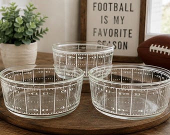 Pode incluir: Três tigelas de vidro transparente com um design impresso em branco que imita as marcações de um campo de futebol americano, incluindo linhas de jardas e números. As tigelas são redondas com bordas lisas e dispostas numa bandeja de madeira.