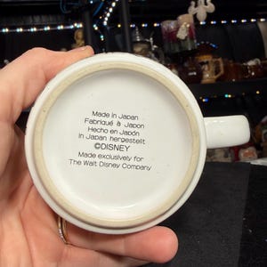 May include: A white ceramic mug with a handle, featuring text that reads "Made in Japan" and "&copy;DISNEY". The mug is made exclusively for The Walt Disney Company. The mug is held by a person.