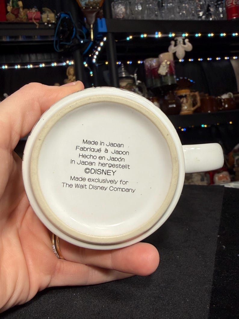 May include: A white ceramic mug with a handle, featuring text that reads "Made in Japan" and "&copy;DISNEY". The mug is made exclusively for The Walt Disney Company. The mug is held by a person.
