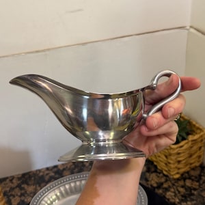 May include: A polished silver-toned gravy boat featuring a curved handle, a wide pouring spout, and a circular pedestal base. The metallic surface reflects light, showcasing a smooth, rounded body designed for serving sauces or gravies.