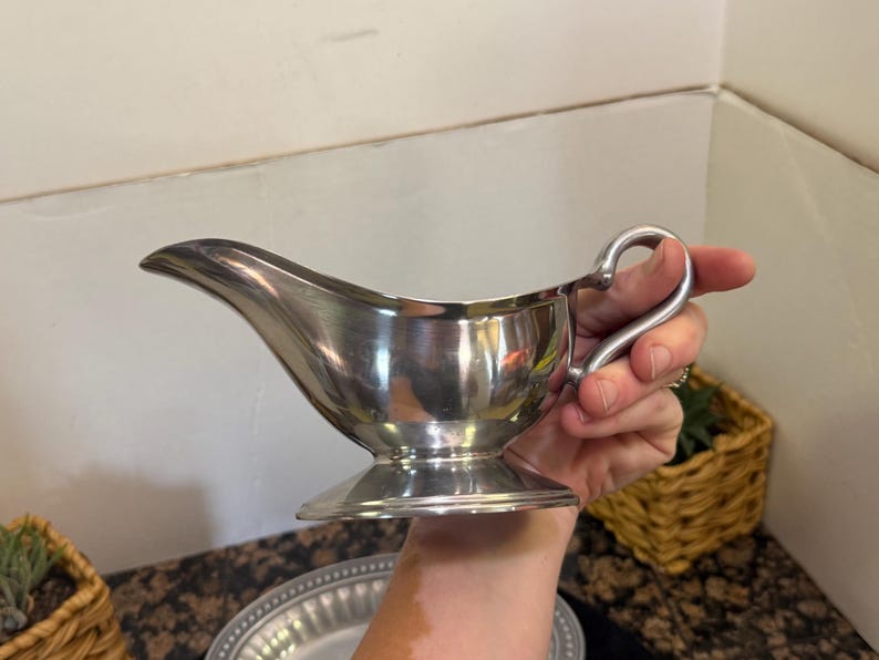 May include: A polished silver-toned gravy boat featuring a curved handle, a wide pouring spout, and a circular pedestal base. The metallic surface reflects light, showcasing a smooth, rounded body designed for serving sauces or gravies.