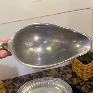 May include: A silver-toned metal gravy boat with a smooth, matte finish and a rounded, teardrop-shaped bowl. The vessel features a single curved handle on one side and a wide, tapered pouring spout, presenting a simple and functional design.