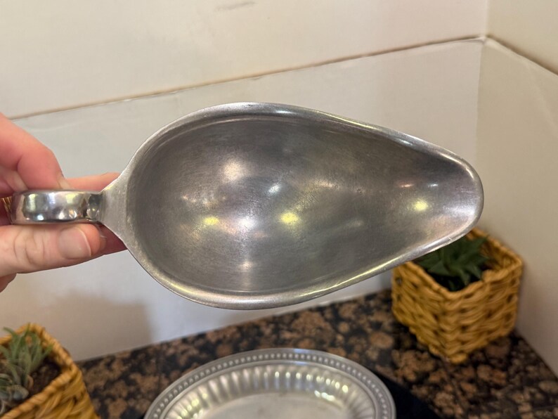 May include: A silver-toned metal gravy boat with a smooth, matte finish and a rounded, teardrop-shaped bowl. The vessel features a single curved handle on one side and a wide, tapered pouring spout, presenting a simple and functional design.
