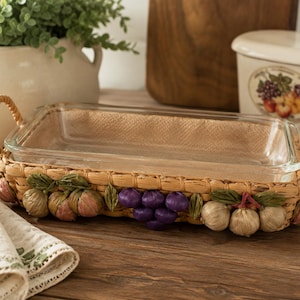 May include: A rectangular clear glass baking dish set inside a woven tan wicker basket holder with two side handles. The basket is decorated with clusters of sculpted, painted fruit including purple grapes, green leaves, and light brown garlic bulbs.