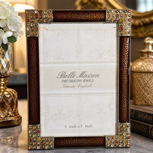 May include: A decorative photo frame with a brown textured border and jeweled corners. The frame is labeled "Belle Maison Decorative Jewels Genuine Crystals" and is sized at 5 inch x 7 inch.
