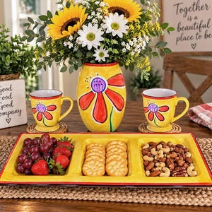May include: A yellow ceramic serving tray with three rectangular compartments containing red grapes, strawberries, round crackers, and mixed nuts. The set includes a matching yellow vase with a painted red flower and two yellow mugs with red flower designs.