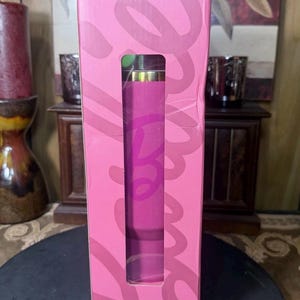 May include: A pink box with a rectangular window revealing a cylindrical object. The box has the word "Love" in a cursive font. The cylindrical object is pink with a gold top. The box is on a dark surface.