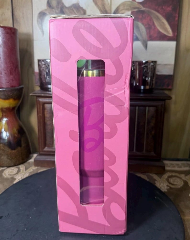May include: A pink box with a rectangular window revealing a cylindrical object. The box has the word "Love" in a cursive font. The cylindrical object is pink with a gold top. The box is on a dark surface.