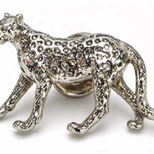 May include: A silver-toned metal lapel pin shaped like a walking leopard. The surface features a textured, raised spot pattern across the body, head, and tail, with a polished, reflective finish and a circular clutch backing visible behind the figure.