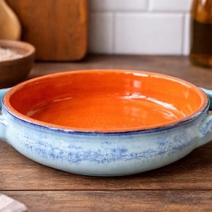 May include: A ceramic serving dish with an orange interior and a light blue exterior. The dish has two handles and a blue rim. The dish is sitting on a wooden surface.