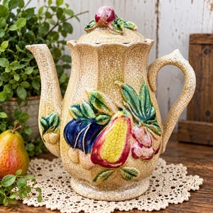 May include: A ceramic teapot with a cream-colored crackle glaze finish, featuring a raised, colorful fruit motif including a yellow pear, blue plum, and red cherries with green leaves. The lid has a small sculpted fruit finial.