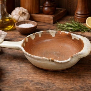 May include: A rustic, handmade ceramic serving dish with a handle. The dish has a cream-colored exterior with a brown interior and a unique, irregular rim. The background includes ingredients like tomatoes, garlic, and rosemary, suggesting a culinary theme.