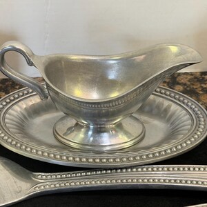May include: A silver-toned metal gravy boat with a curved handle and a matching oval underplate featuring a beaded rim. The gravy boat has a pedestal base and a smooth interior, while the underplate displays a fluted design and a beaded edge.