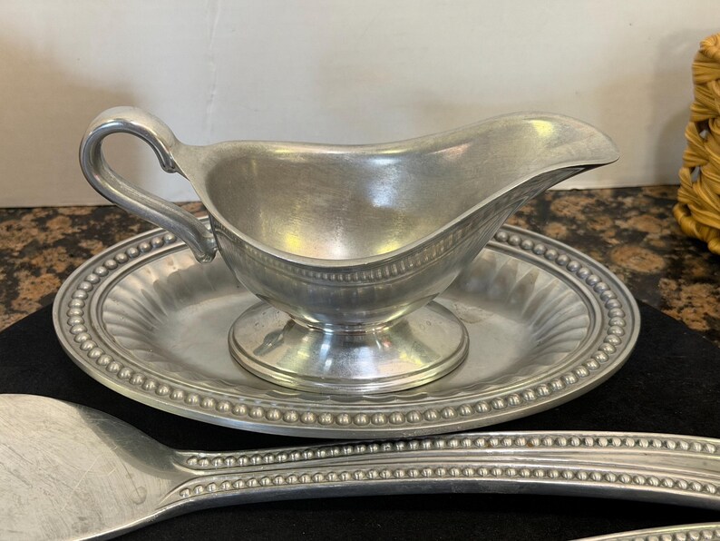 May include: A silver-toned metal gravy boat with a curved handle and a matching oval underplate featuring a beaded rim. The gravy boat has a pedestal base and a smooth interior, while the underplate displays a fluted design and a beaded edge.