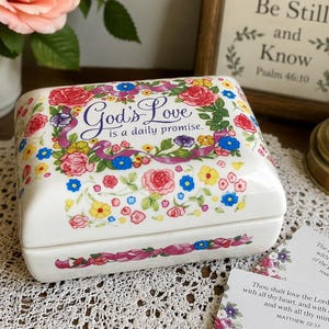 May include: A white ceramic trinket box with a floral design featuring red, pink, blue, and yellow flowers. The box is adorned with the text "God's Love is a daily promise." A decorative item for storing small items.