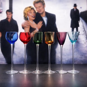 May include: A set of six colorful stemmed glasses. Each glass has a different colored bowl: blue, red, green, amber, purple, and teal. The glasses are arranged in a row, with a blurred background.