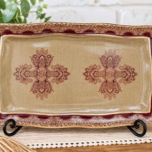 May include: A rectangular ceramic tray featuring a tan center with two symmetrical dark red and gold ornate paisley patterns. The tray has a scalloped edge with a deep red border and a detailed gold filigree design along the entire rim.