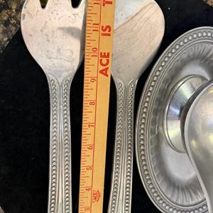 May include: A set of silver-toned metal salad servers featuring a beaded handle design, positioned next to a wooden ruler that shows a length of approximately 13 inches. A matching silver-toned metal gravy boat with a beaded rim is also visible.