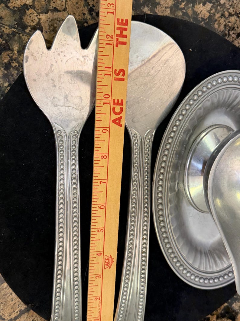 May include: A set of silver-toned metal salad servers featuring a beaded handle design, positioned next to a wooden ruler that shows a length of approximately 13 inches. A matching silver-toned metal gravy boat with a beaded rim is also visible.