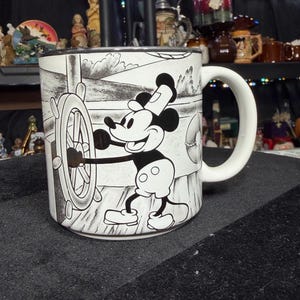 May include: A white ceramic mug with a black and white illustration of Mickey Mouse steering a ship's wheel. The mug has a curved white handle and a black rim. The design shows Mickey Mouse at the helm of a ship.