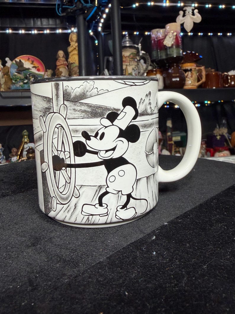 May include: A white ceramic mug with a black and white illustration of Mickey Mouse steering a ship's wheel. The mug has a curved white handle and a black rim. The design shows Mickey Mouse at the helm of a ship.