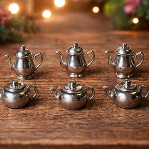 May include: A collection of six miniature silver-toned metal teapots arranged in two rows. The top row features three taller, upright teapots, while the bottom row contains three shorter, rounded teapots, all with lids, handles, and spouts.