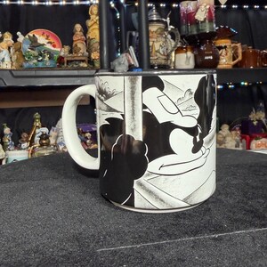 May include: A white ceramic mug with a black and white cartoon illustration. The mug has a curved handle and a design that wraps around the body. Other decorative items are visible in the background.