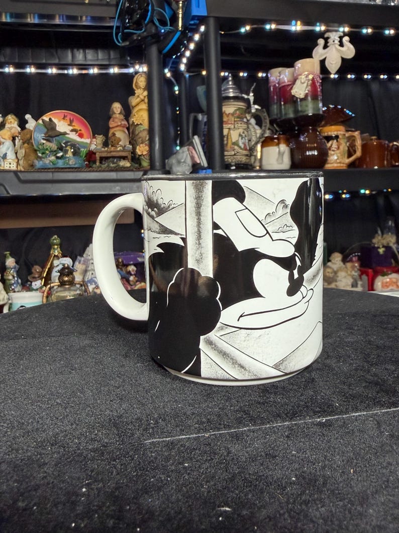 May include: A white ceramic mug with a black and white cartoon illustration. The mug has a curved handle and a design that wraps around the body. Other decorative items are visible in the background.
