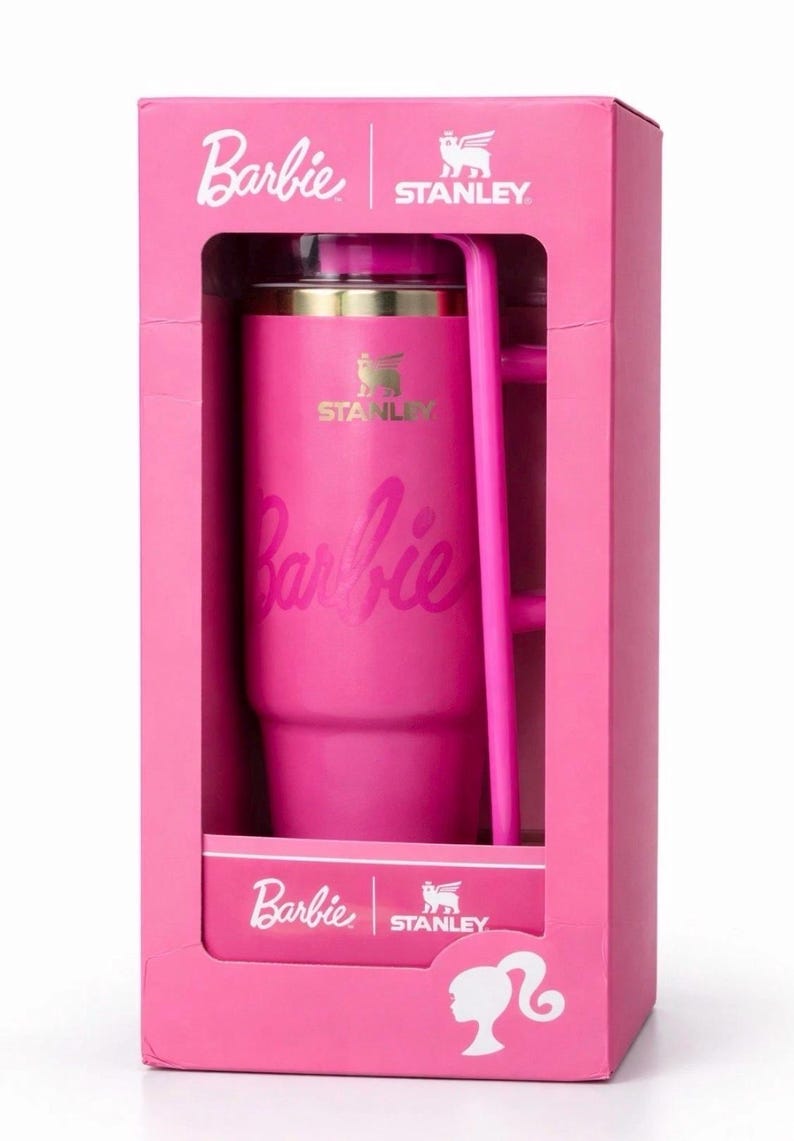 May include: A pink Stanley tumbler with the Barbie logo in a pink box. The tumbler has a handle and a gold rim. The box also features the Barbie and Stanley logos.