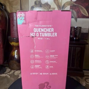 May include: A pink cardboard box for a Flowstate Quencher H2.0 Tumbler. The box features the Barbie 65 logo and product details, including 40 oz capacity. The box also lists features like stainless steel and double-wall vacuum insulation.
