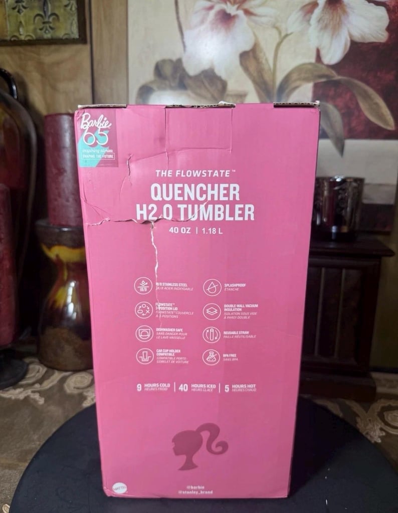 May include: A pink cardboard box for a Flowstate Quencher H2.0 Tumbler. The box features the Barbie 65 logo and product details, including 40 oz capacity. The box also lists features like stainless steel and double-wall vacuum insulation.