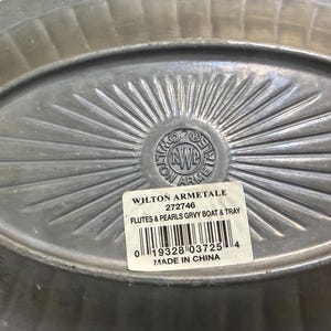 May include: An oval-shaped Wilton Armetale metal tray with a fluted rim and a sunburst pattern radiating from the center. A white rectangular sticker is affixed to the surface, displaying the text WILTON ARMETALE 272746 FLUTES & PEARLS GRVY BOAT & TRAY.