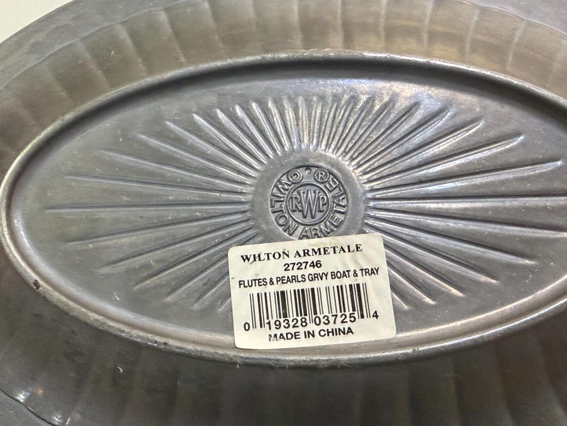 May include: An oval-shaped Wilton Armetale metal tray with a fluted rim and a sunburst pattern radiating from the center. A white rectangular sticker is affixed to the surface, displaying the text WILTON ARMETALE 272746 FLUTES & PEARLS GRVY BOAT & TRAY.