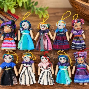 May include: Ten small handmade dolls with embroidered facial features and yarn hair, each wearing a woven fabric dress in various colors and patterns. Each doll has a yellow or red string loop attached to the top of the head for hanging.