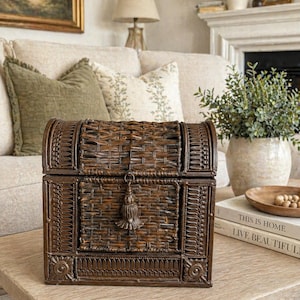 May include: A dark brown woven chest with a curved top and a rectangular front. The chest has a decorative tassel and is sitting on a light-colored wooden table. The chest is in a living room setting.
