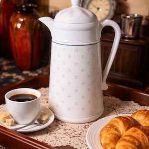 May include: A white ceramic coffee pot with a lid, handle, and spout, decorated with small blue floral patterns. A cup of coffee, sugar cubes, and croissants are on a wooden tray, suggesting a breakfast or brunch setting.