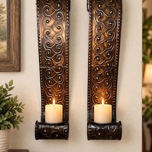 May include: A pair of tall, dark bronze-colored metal wall sconces featuring an intricate embossed scrollwork pattern and a studded border. Each sconce has a curved base holding a single white pillar candle with a lit flame.