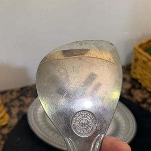 May include: A silver-toned metal serving spoon with a wide, rounded bowl. The surface shows signs of wear and light scratches. A circular hallmark is stamped near the handle, featuring the text RWP and WILTON ARMETALE.