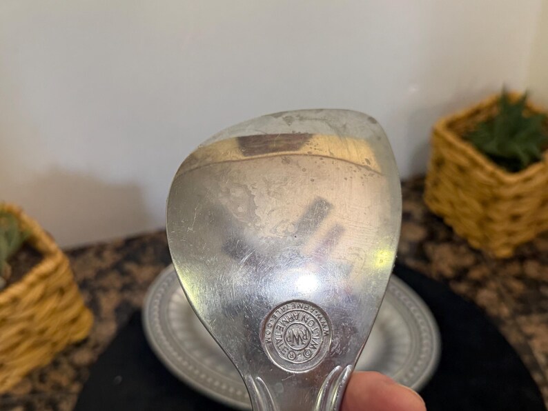 May include: A silver-toned metal serving spoon with a wide, rounded bowl. The surface shows signs of wear and light scratches. A circular hallmark is stamped near the handle, featuring the text RWP and WILTON ARMETALE.