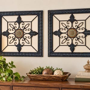 May include: Two square wall art pieces featuring black metal scrollwork frames with ornate edges. Each piece contains a central circular medallion with intricate patterns in shades of green, brown, and cream, set against a light background.